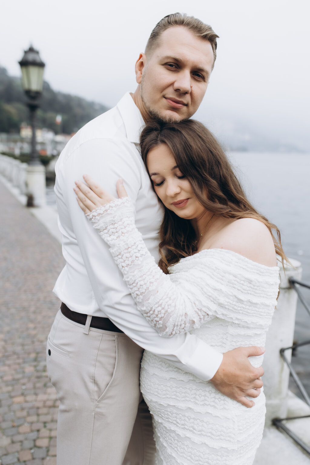 her rests against him in a tender embrace on the Como lakefront, her eyes closed, the misty lake behind them, on Lake Como.
