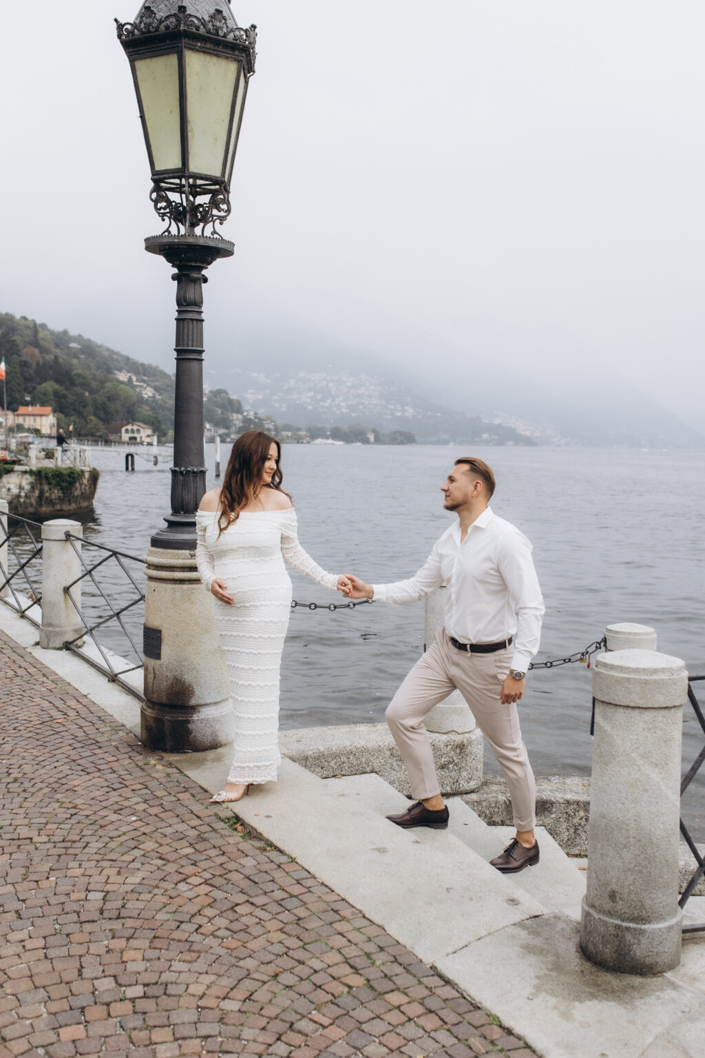 her holds his hand beside an ornate wrought-iron lamppost on the Como lakefront promenade, the misty hills behind them, on Lake Como.