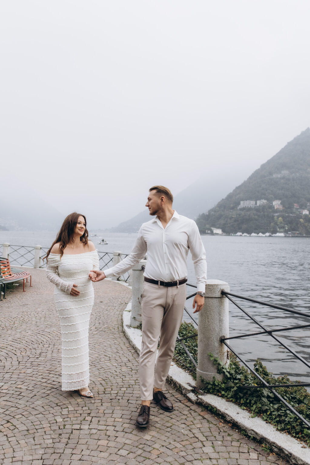 A newly engaged couple walk hand in hand along the railed lakefront promenade of Como, the misty hills behind them, on Lake Como.