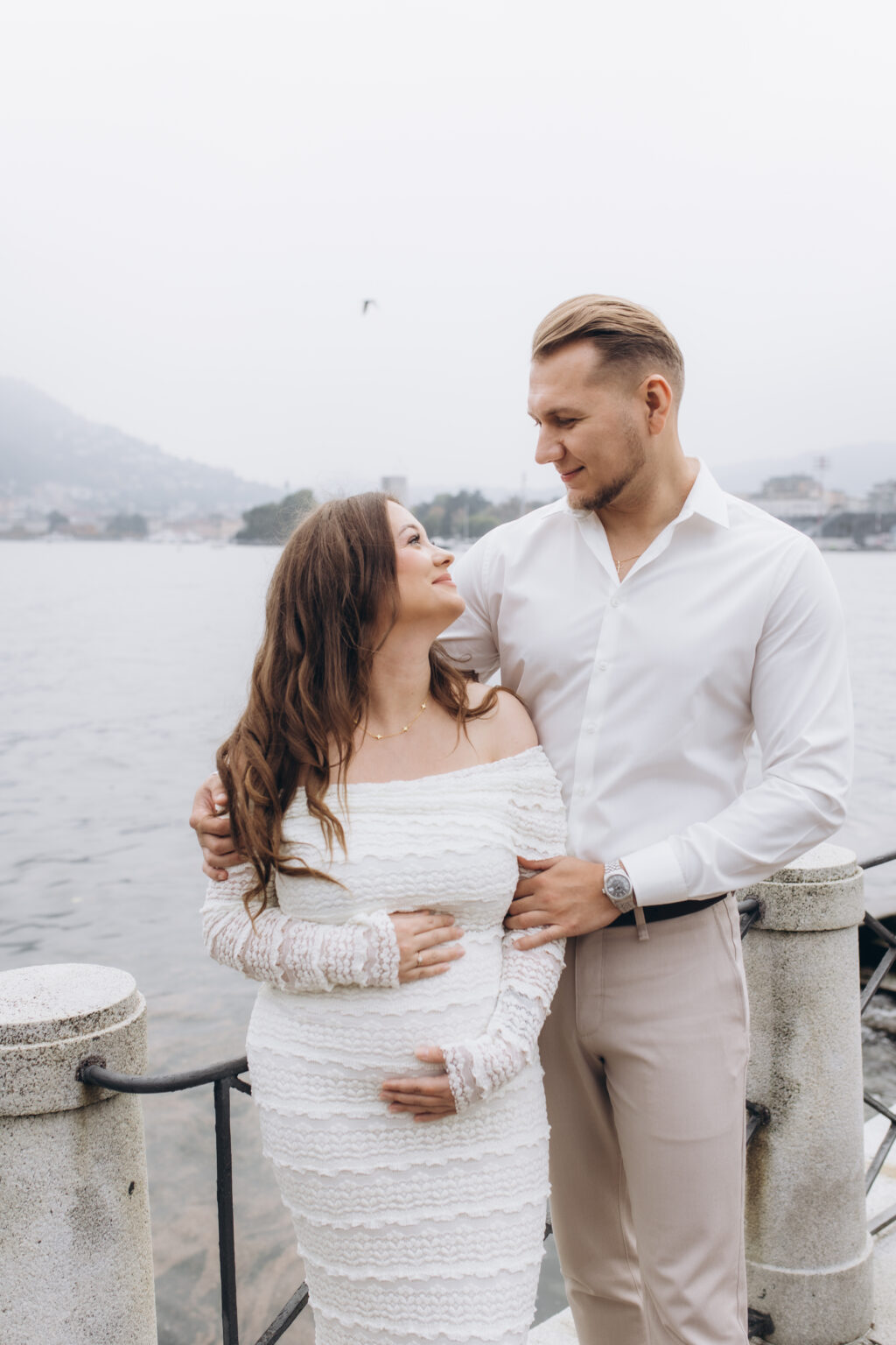 A newly engaged couple hold each other by the lakefront railing of Como, both hands resting on her bump, the misty hills behind them, on Lake Como.