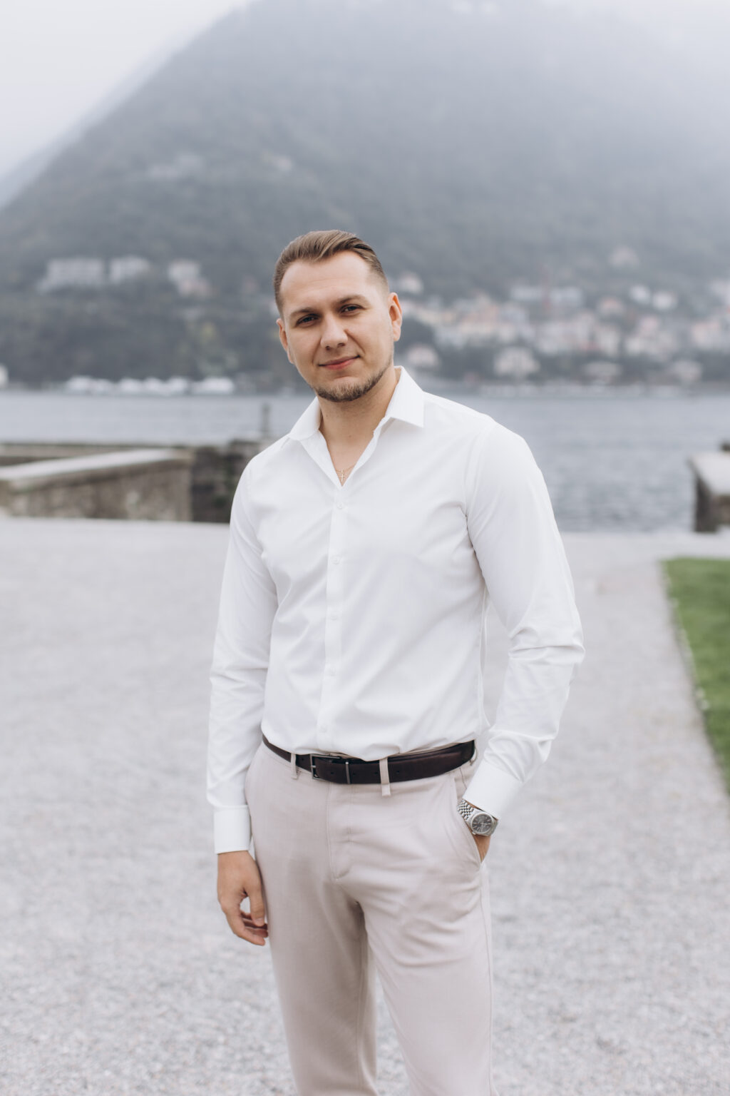He stands alone in a white shirt with his hand in his pocket on the lakefront of Villa Olmo, the misty Como mountains behind him, on Lake Como.