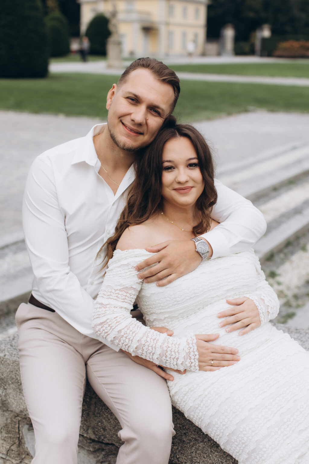 A newly engaged couple sit close, both smiling at the camera with hands resting on her bump, the ochre Villa Olmo villa behind them, on Lake Como.