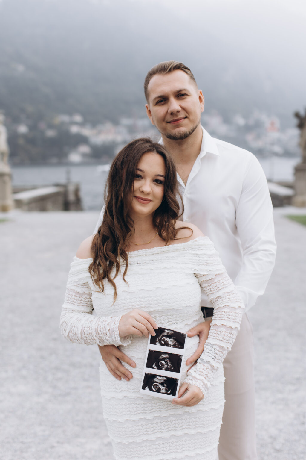 She in a white lace off-shoulder dress holds an ultrasound scan over her baby bump as he stands behind her, before the misty lake at Villa Olmo, on Lake Como.