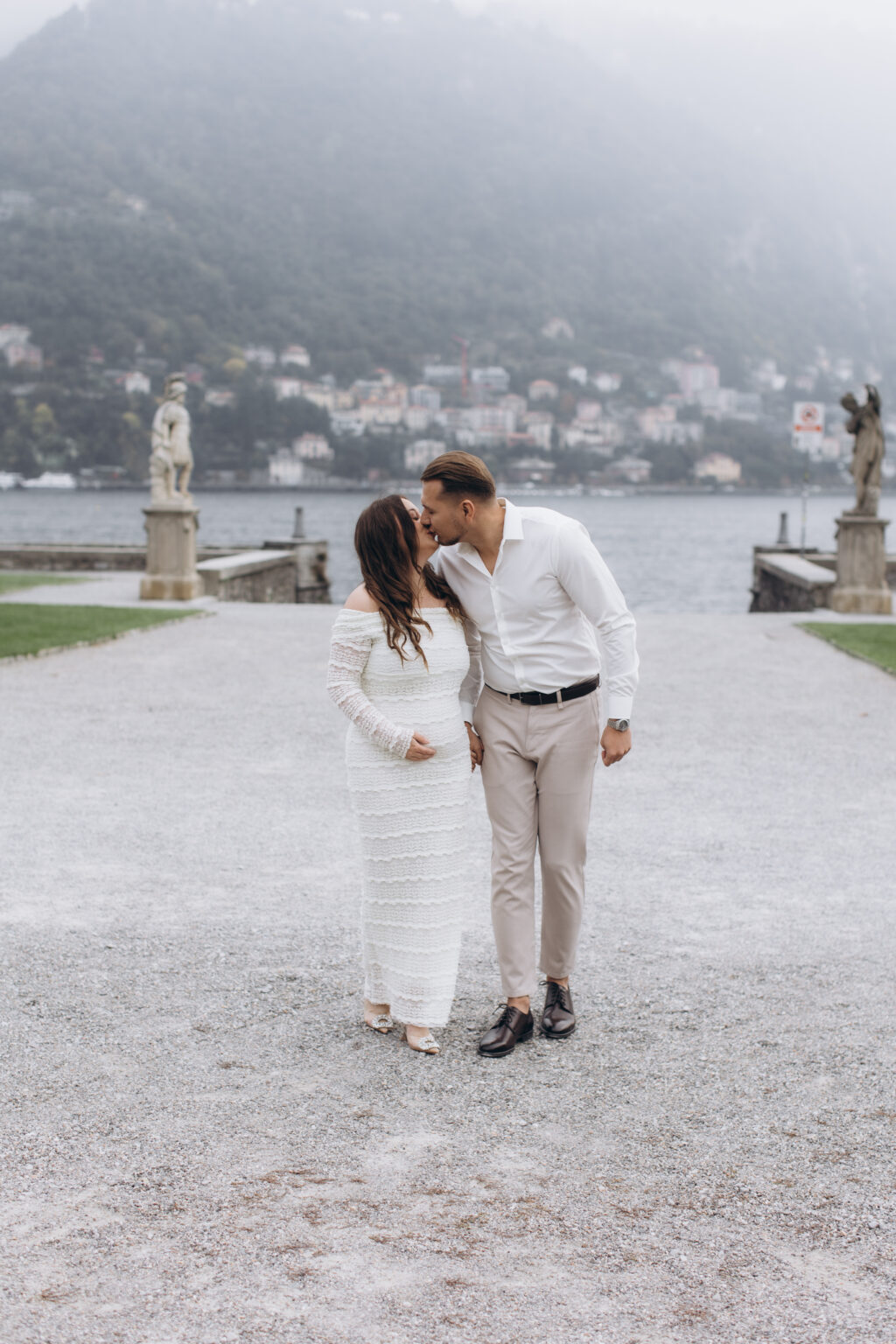 The couple share a kiss on the gravel terrace of Villa Olmo, flanked by neoclassical statues, with the misty lake behind them, on Lake Como.