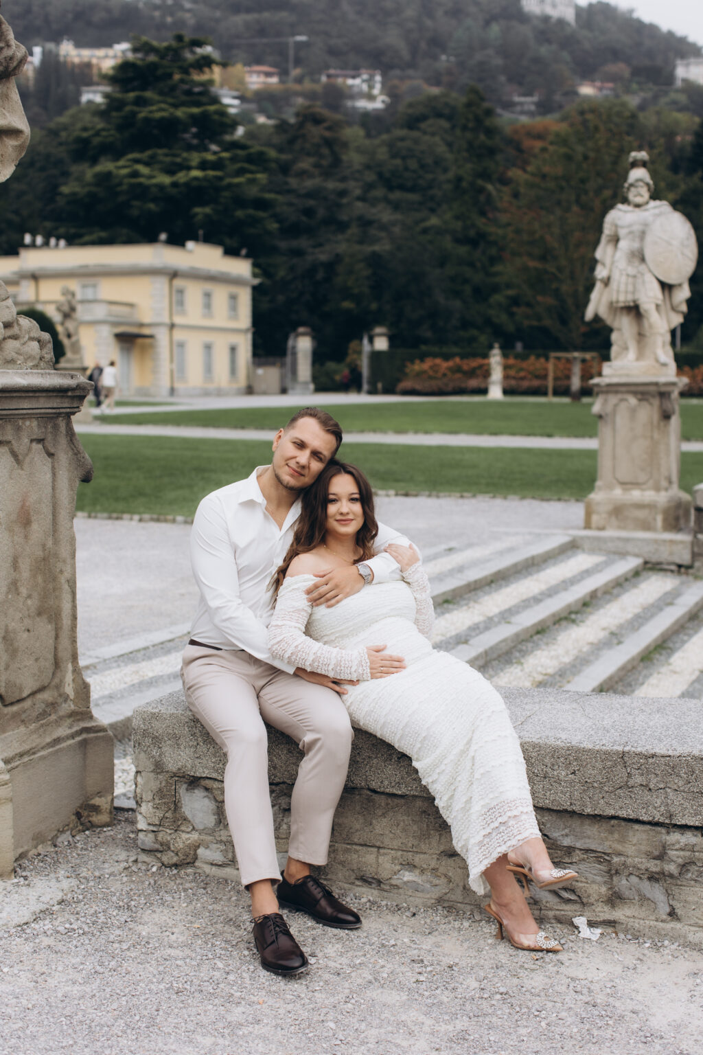 A newly engaged couple sit close on a stone ledge, a warrior statue and an ochre villa in the park of Villa Olmo behind them, on Lake Como.
