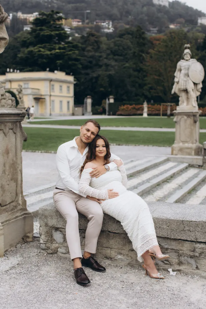 A newly engaged couple sit close on a stone ledge, a warrior statue and an ochre villa in the park of Villa Olmo behind them, on Lake Como.