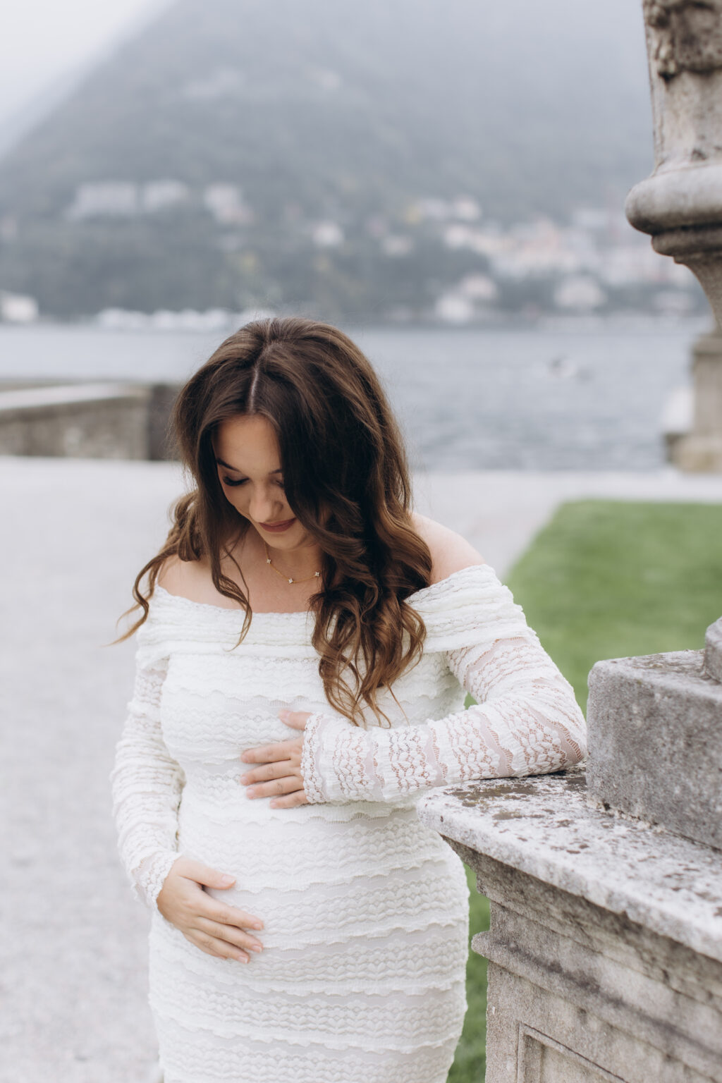 She cradles her baby bump with both hands, looking down, beside a stone baluster in the gardens of Villa Olmo, the misty lake behind her, on Lake Como.