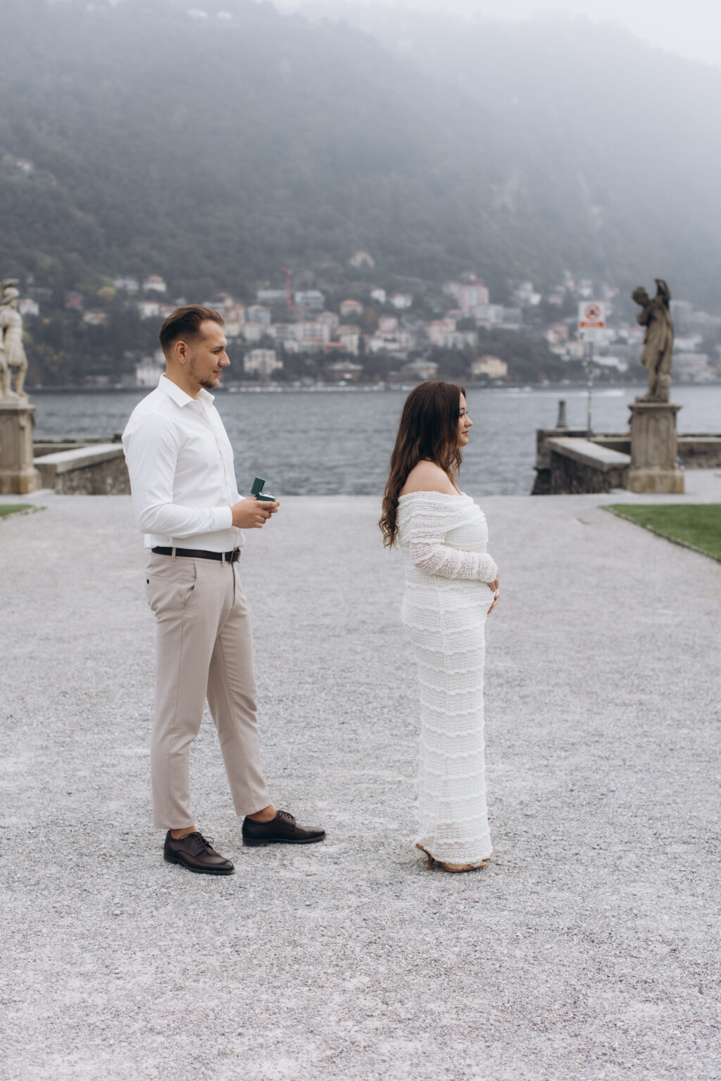 He faces his pregnant partner across the gravel terrace, ring box in hand, neoclassical statues and the misty lake behind them, in the gardens of Villa Olmo on Lake Como.