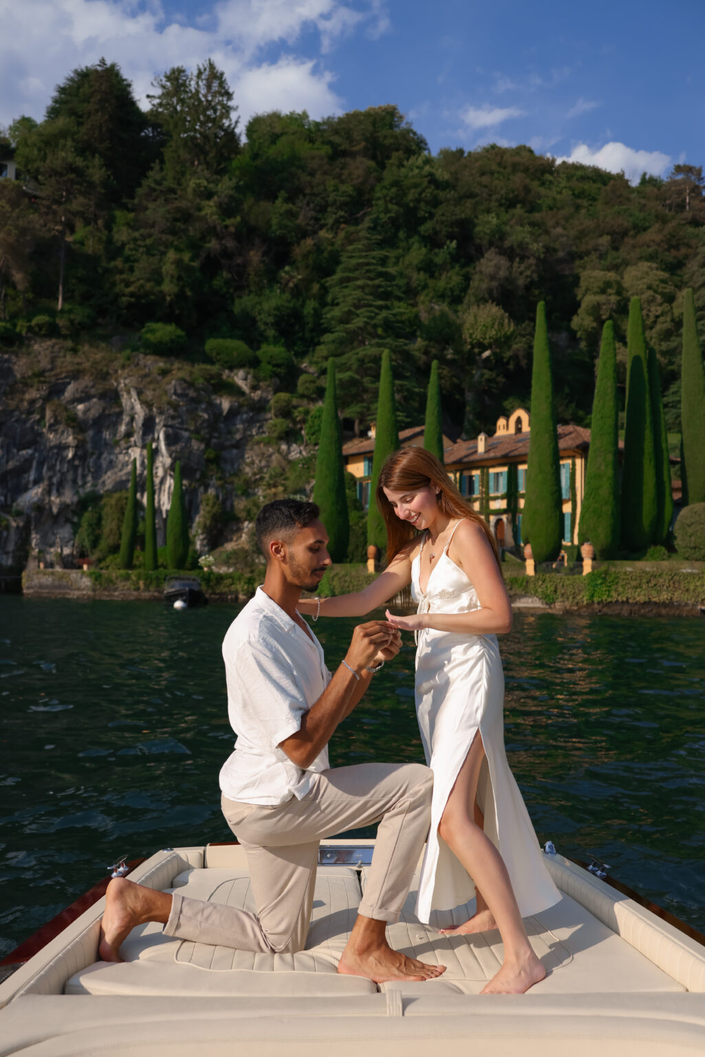A kneeling man slides an engagement ring onto a smiling woman's finger on the bow of a boat, before an ochre villa framed by cypresses on Lake Como.