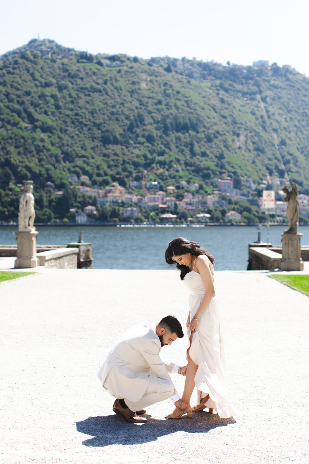 Man kneeling to adjust his partner's sandal on a statue-flanked villa terrace above the water on Lake Como, the town of Como behind them