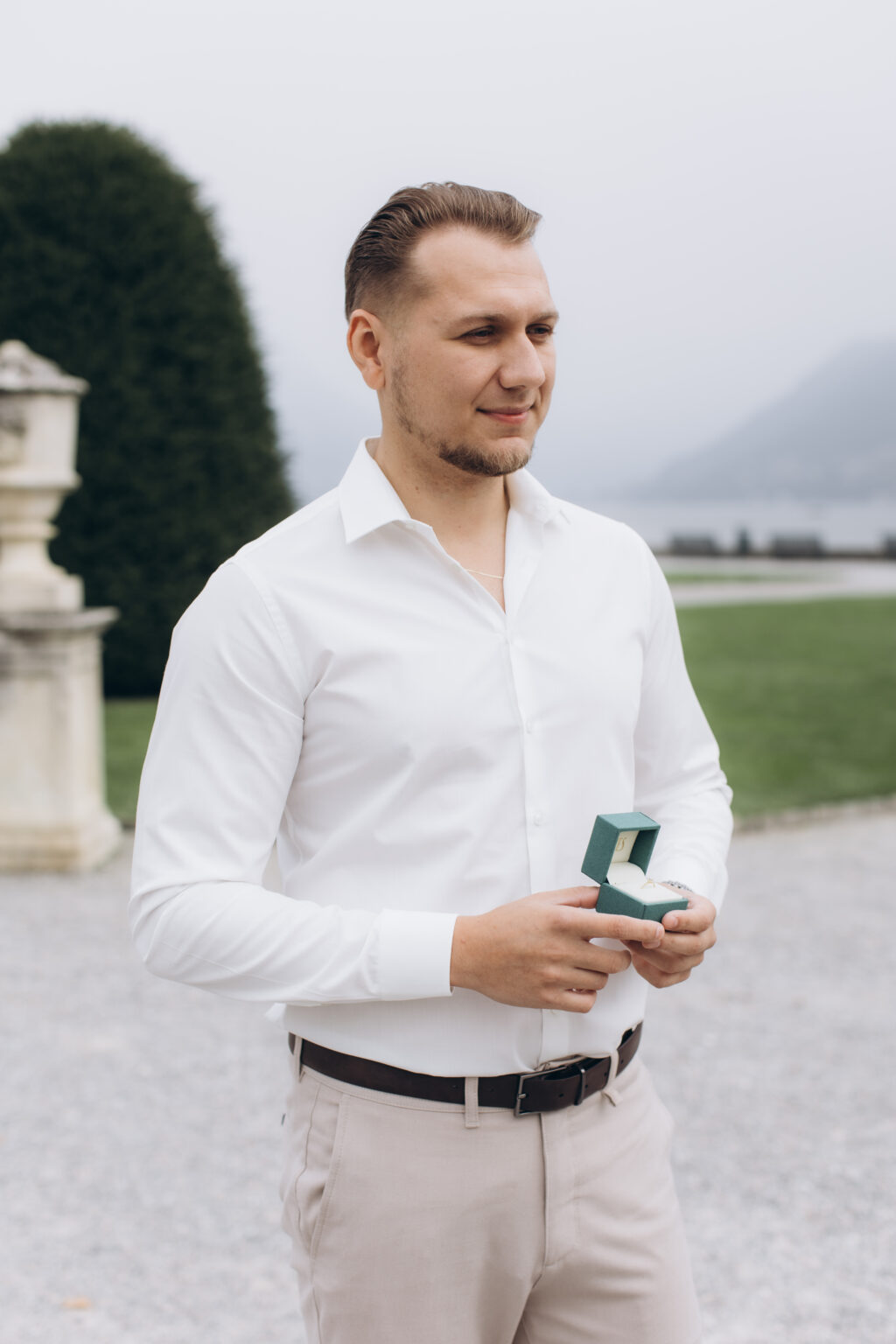 He holds an open green ring box, gazing off, beside a garden urn and hedge in the park of Villa Olmo, on Lake Como.