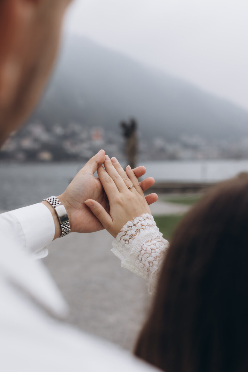 A close-up of her hand resting on his, her new engagement ring visible, with a couple embracing in the misty distance at Villa Olmo, on Lake Como.