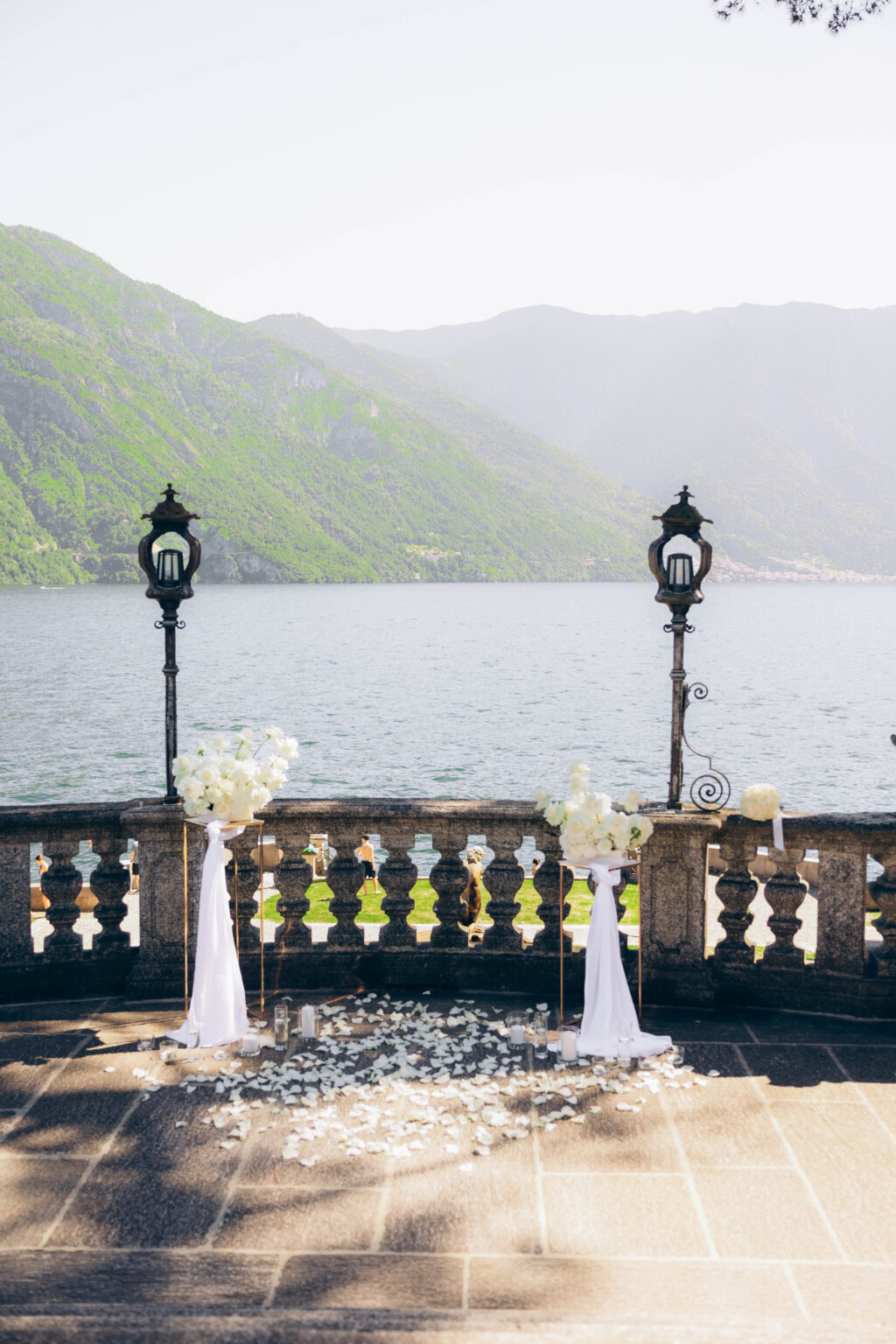 Proposal setup on a balustraded terrace in Parco Teresio Olivelli with white roses on gold stands, chiffon drapes, candles, scattered petals, and two antique lanterns overlooking Lake Como