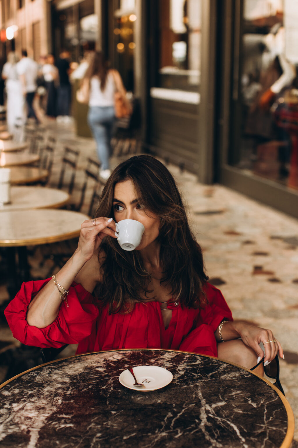 Woman in a red off-shoulder dress sips from an espresso cup at a marble table on a Milan café terrace.