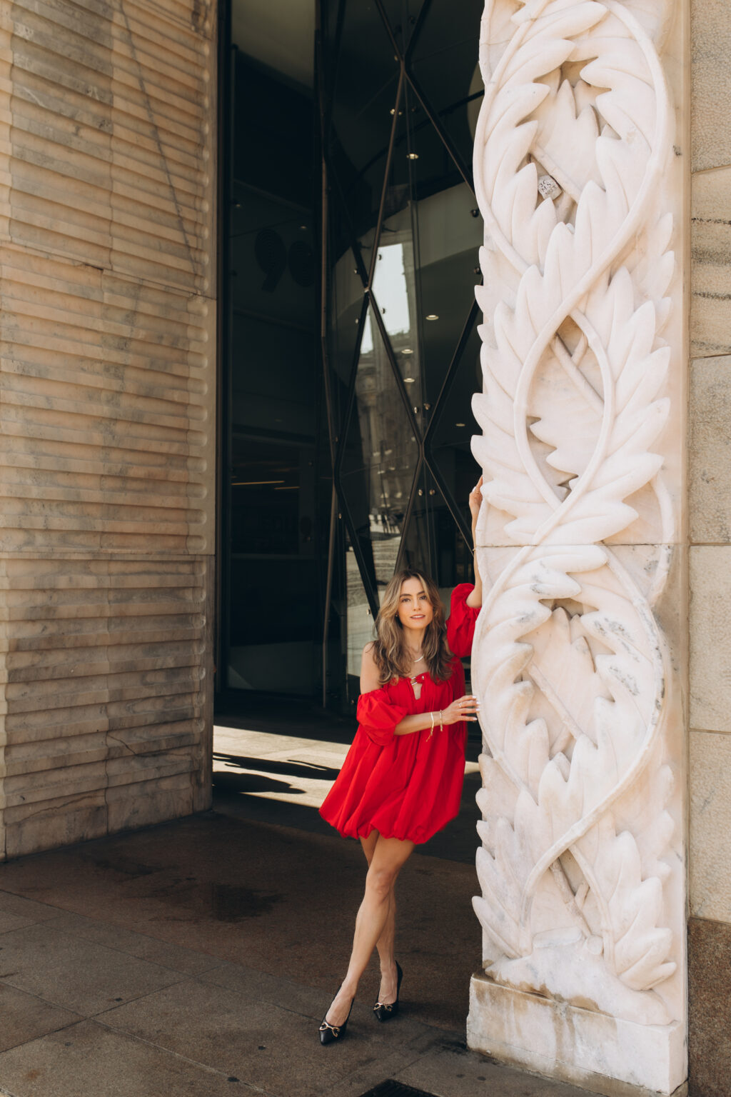 Woman in a red dress leans against a carved marble pilaster near a modern glass facade by Piazza del Duomo in Milan.