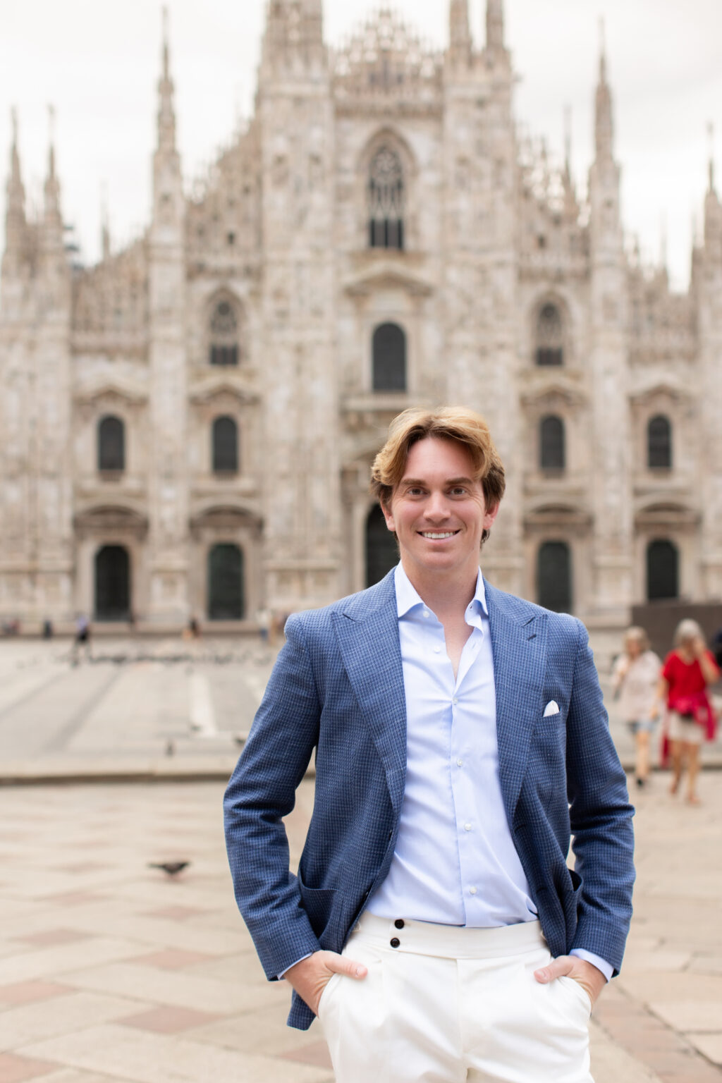 A smiling man in a houndstooth blue jacket standing with hands in pockets in front of the Duomo di Milano in Milan.