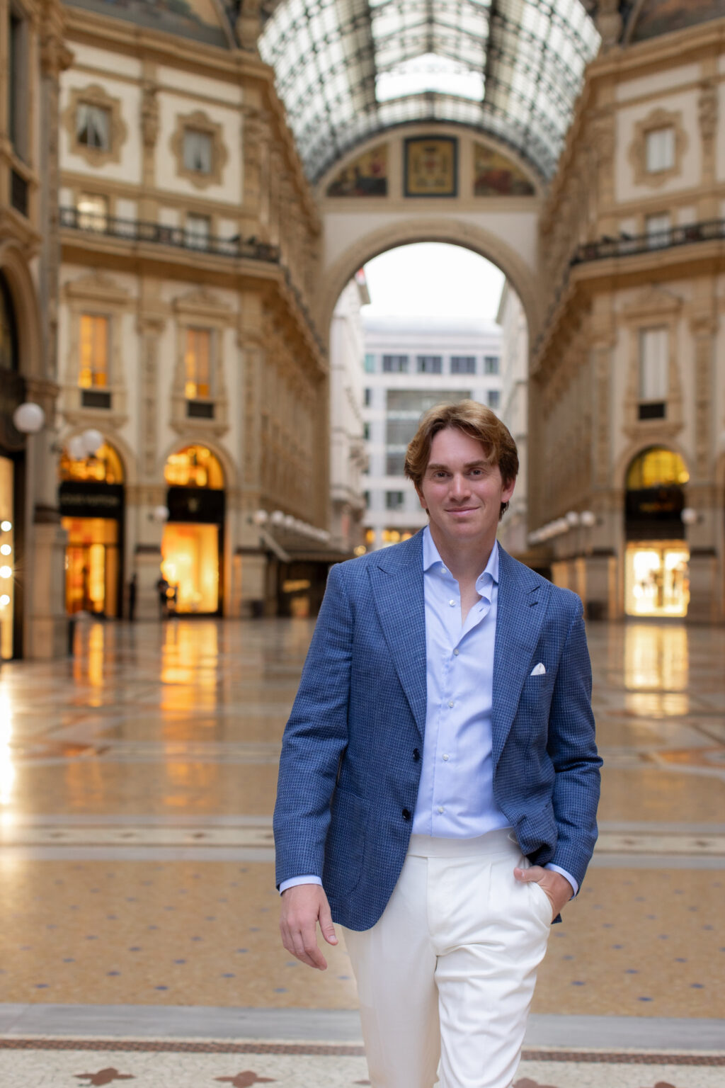 A man in a blue houndstooth jacket walking toward the camera in the Galleria Vittorio Emanuele II, the glass dome behind him.