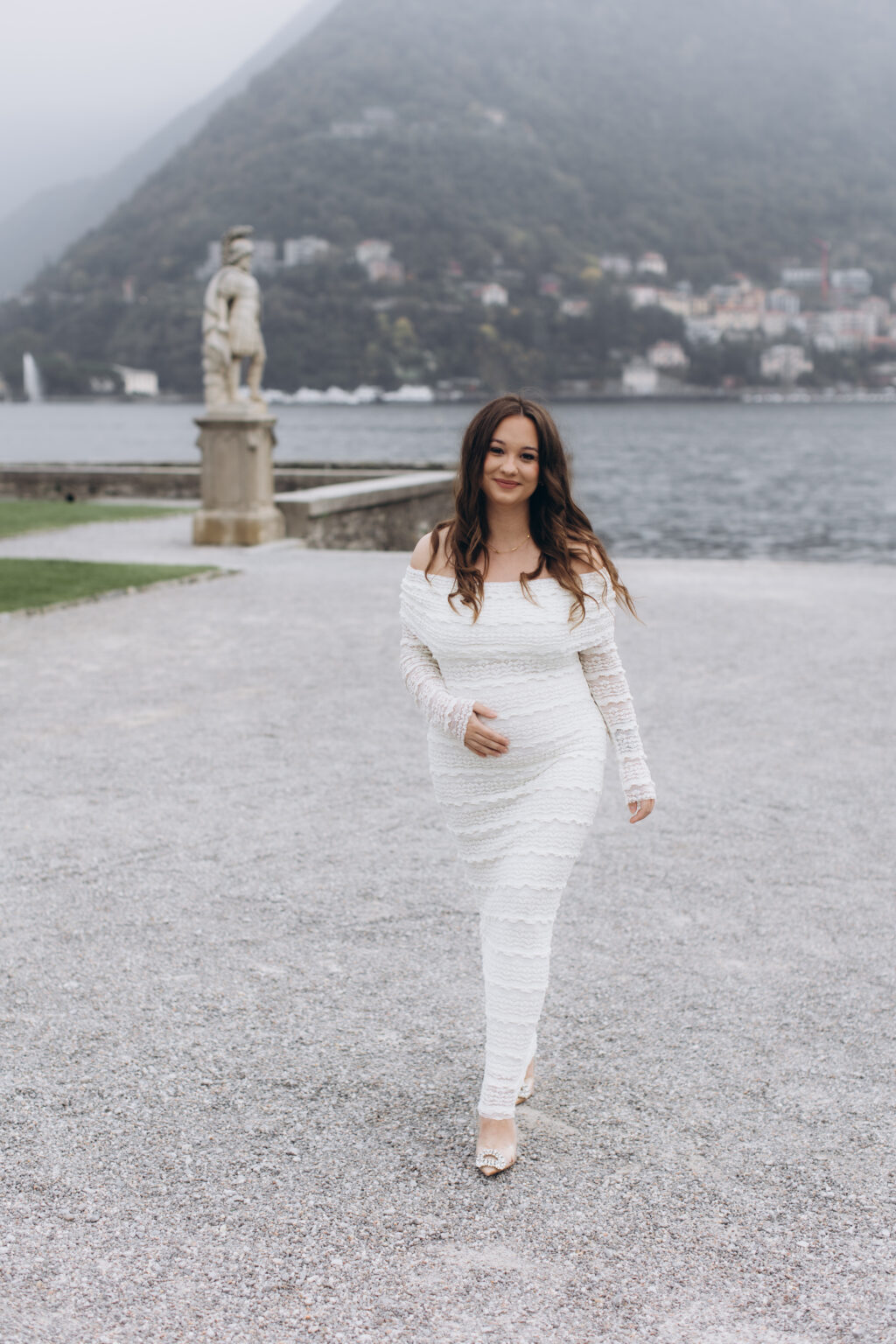 She walks toward the camera cradling her bump, a classical warrior statue and the misty hills of Como behind her, in the gardens of Villa Olmo on Lake Como.