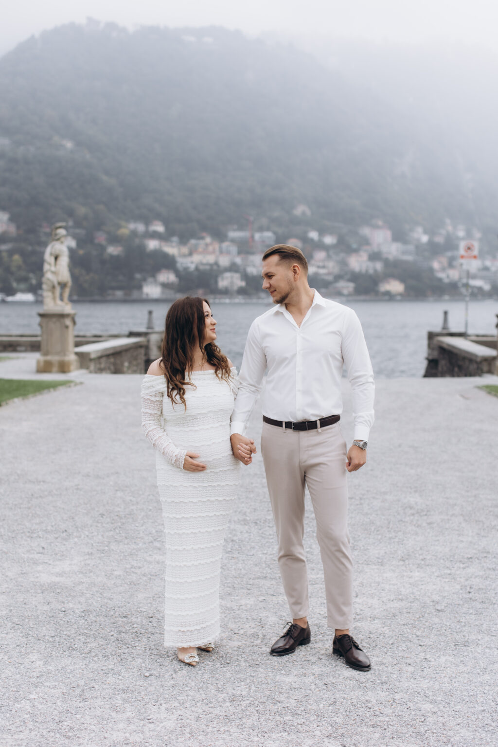 She, one hand on her bump, holds hands with him on the gravel terrace of Villa Olmo, a classical statue and the misty hills of Como behind them, on Lake Como.