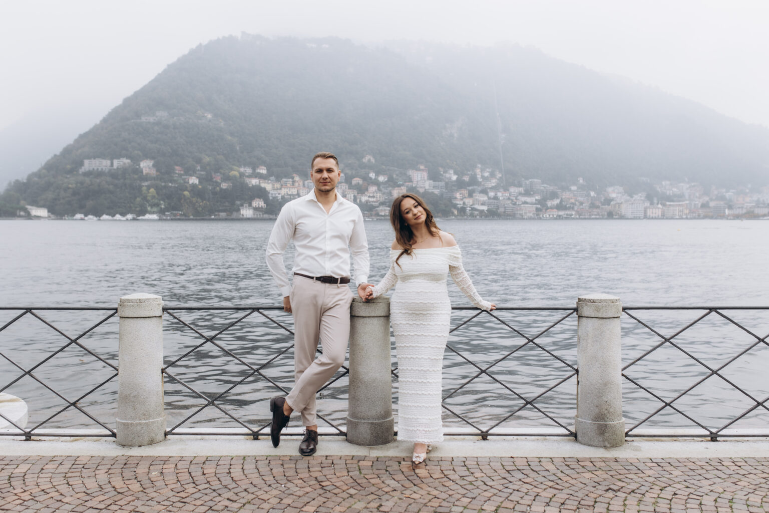 A newly engaged couple lean against the lakefront railing of Como, holding hands, the misty hills and town behind them, on Lake Como.