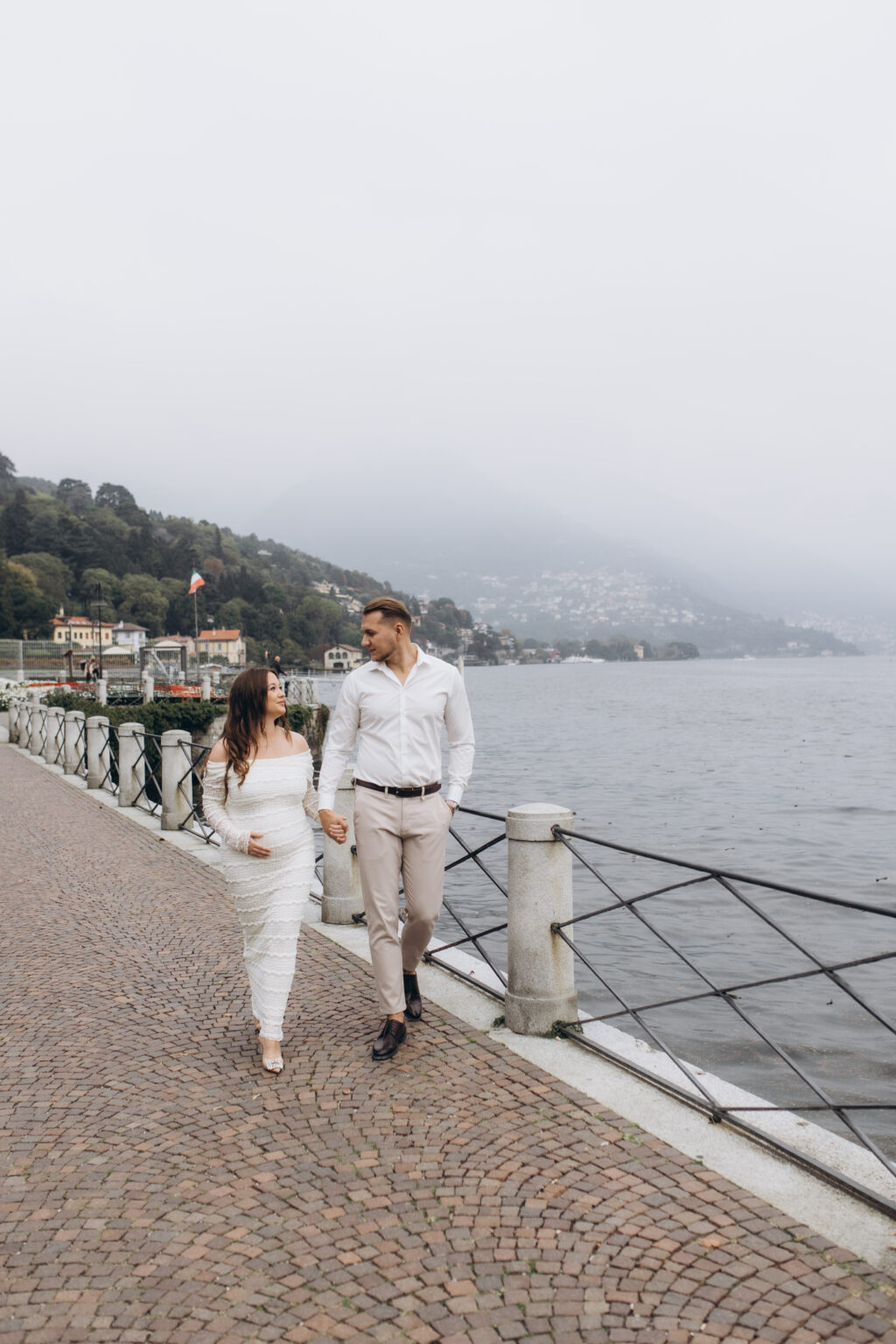 A newly engaged couple walk hand in hand along the Como lakefront promenade past an antique lamppost, the misty lake and hills behind them, on Lake Como.