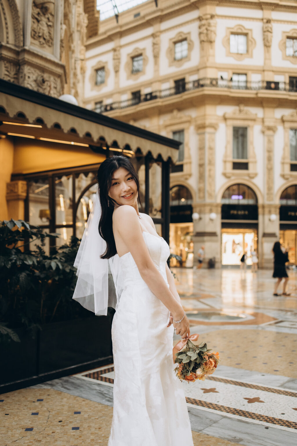Bride-to-be in a white gown and veil looking over her shoulder with her bouquet, the Prada storefront behind her in the Galleria in Milan