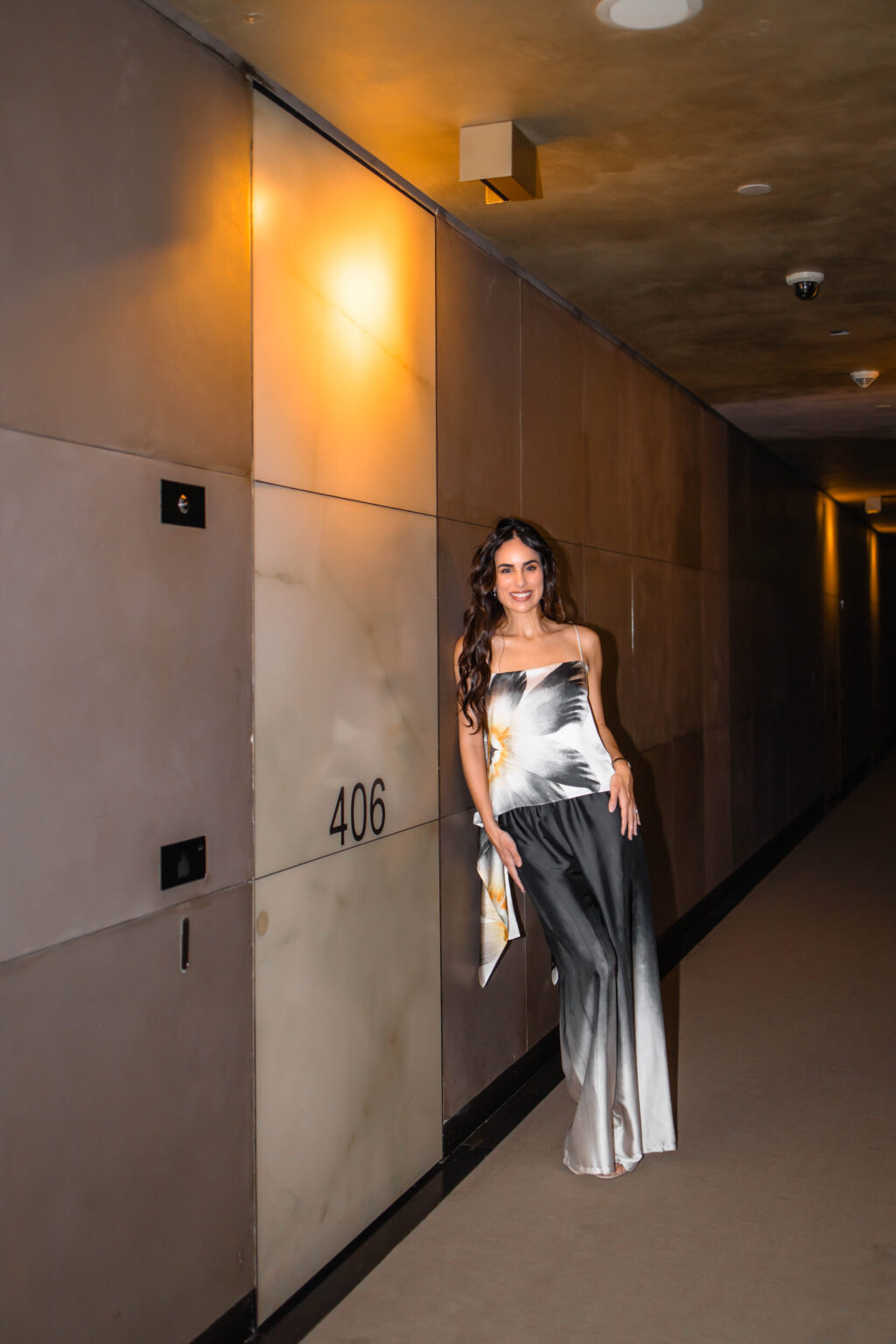 She in a floral silk top and silver-to-black silk trousers leans against the onyx-clad corridor wall beside room 406 at the Armani Hotel Milano.