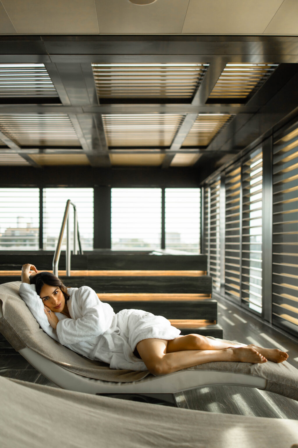 She reclines on a spa lounger in a white robe before the louvered windows and city view of the Armani/SPA at the Armani Hotel Milano.