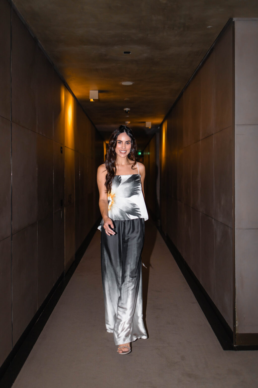 She walks toward the camera in a floral silk top and silver silk trousers along a dark corridor at the Armani Hotel Milano.