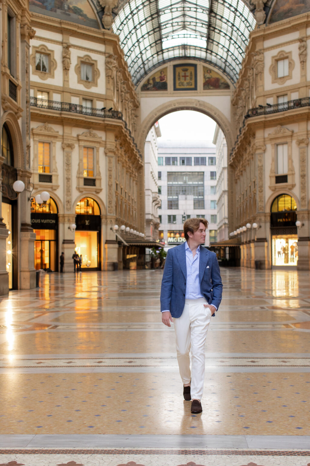 A man in a blue blazer walking across the mosaic floor of the Galleria Vittorio Emanuele II past luxury boutiques in Milan.