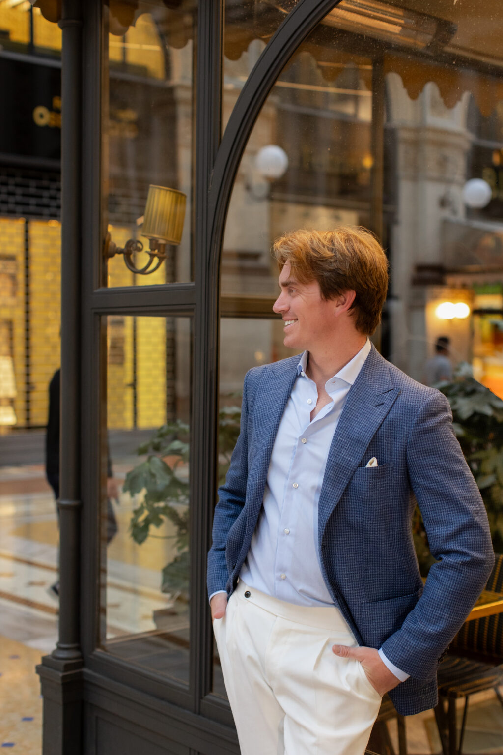 A man in a blue blazer leaning beside an ornate arched cafe window with warm lamplight, inside a historic arcade in central Milan.