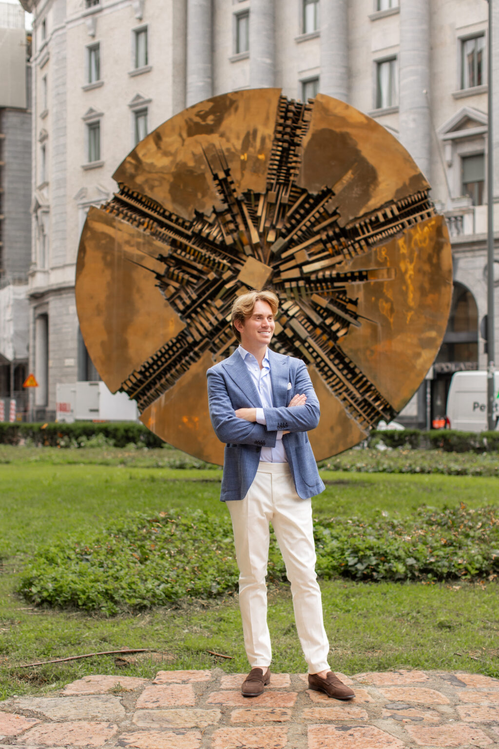 A man in a blue blazer standing with arms crossed in front of Arnaldo Pomodoro's golden Grande Disco sculpture in Piazza Meda, Milan.