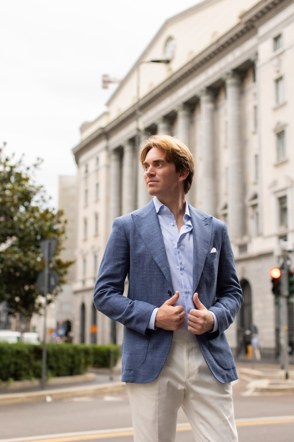 A man in a blue jacket adjusting his lapels in front of a grand neoclassical colonnaded building in central Milan.