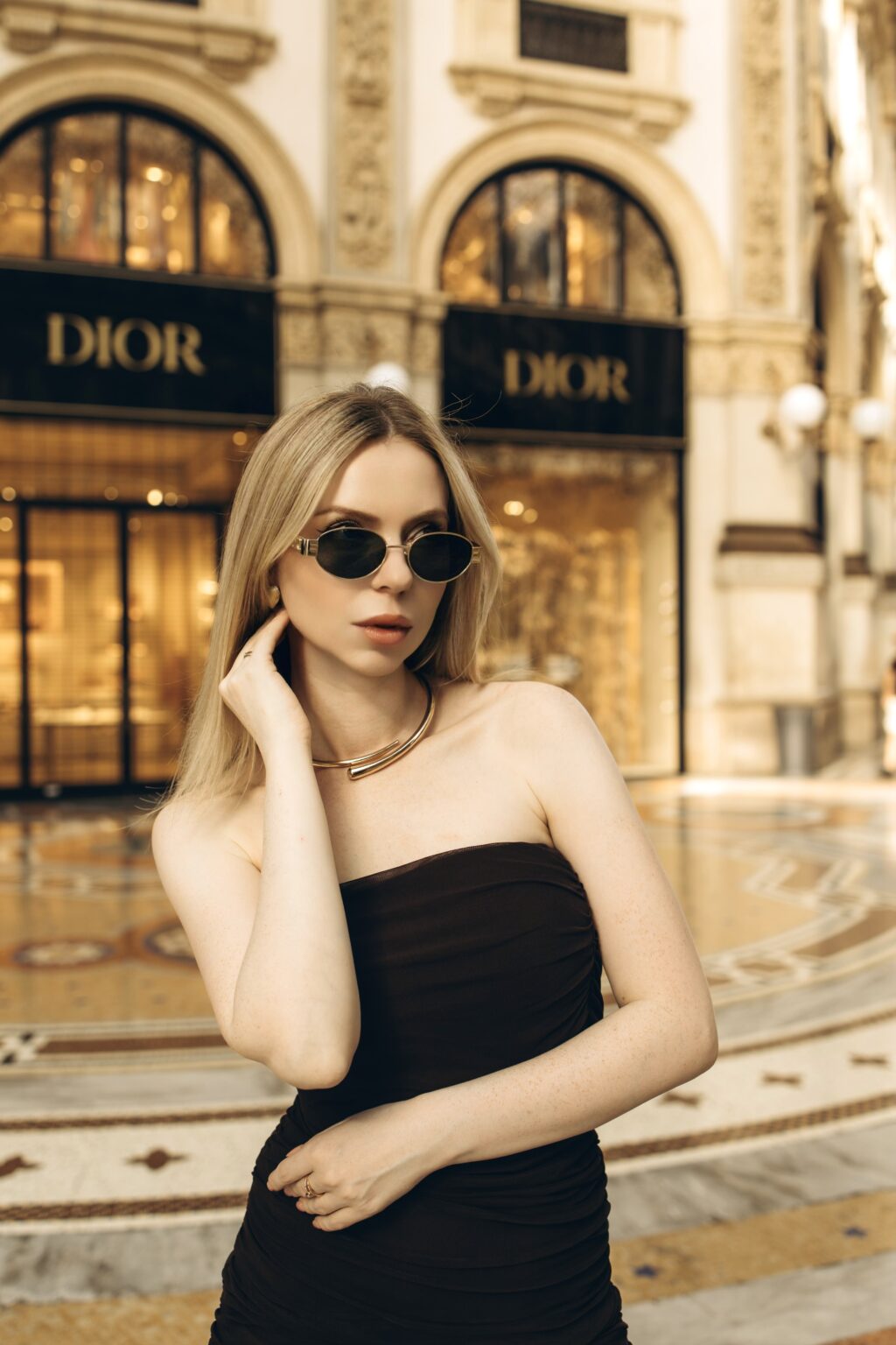 Close-up of a blonde woman in oval sunglasses and a gold collar necklace, hand at her ear, with glowing boutique windows of Galleria Vittorio Emanuele II blurred behind
