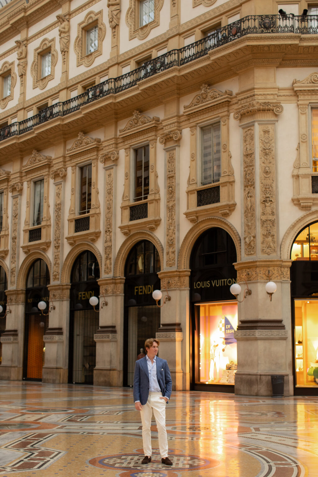 A man standing small against the ornate boutique facade of the Galleria Vittorio Emanuele II, with Louis Vuitton and Fendi shopfronts in Milan.