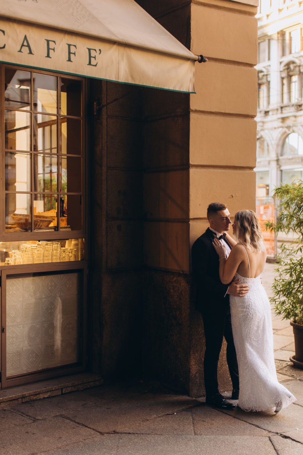 The couple lean close in a near-kiss beside the lit window of a historic caffè near Piazza del Duomo, warm golden light spilling over them.