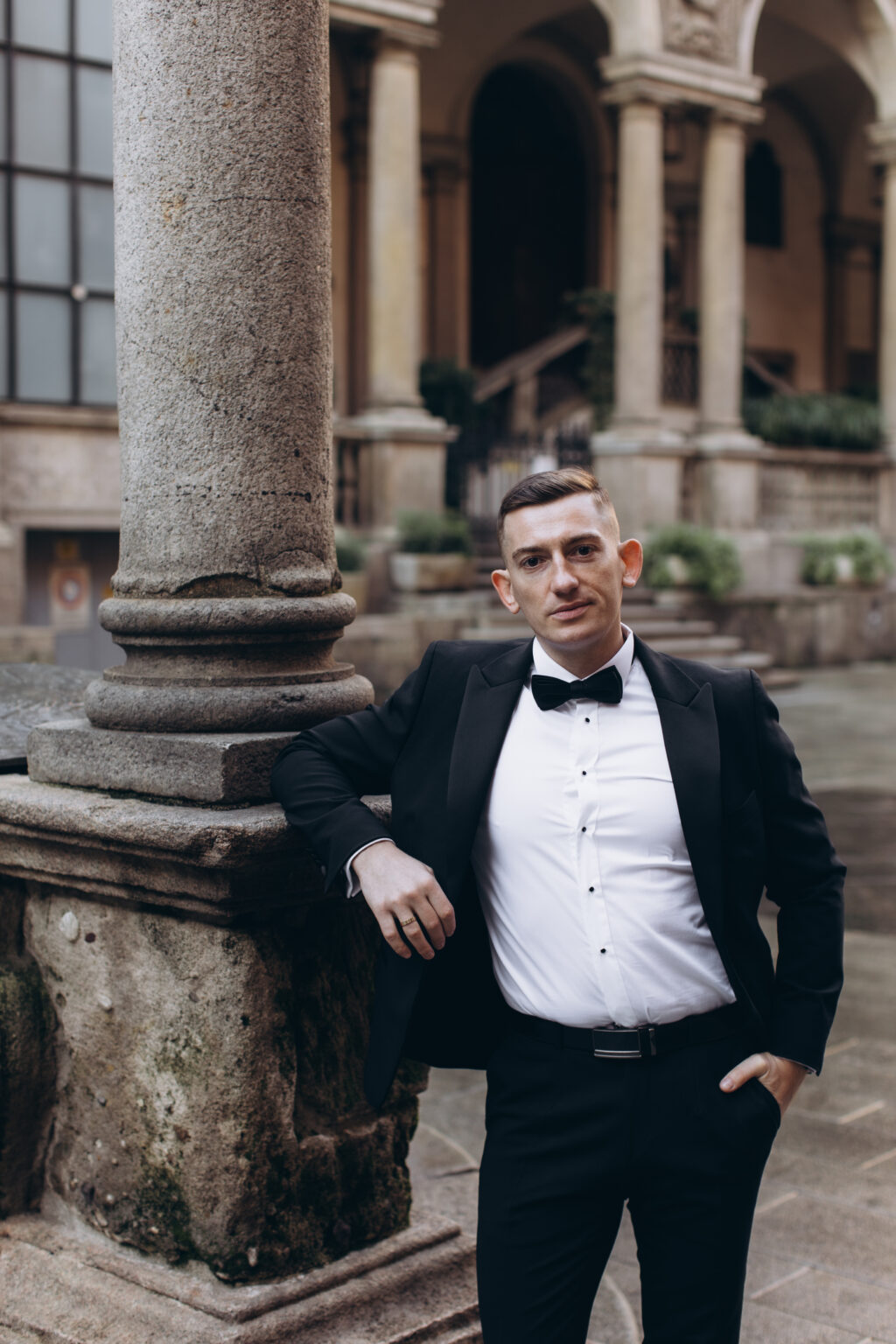 He leans against a granite column in a historic brick-arcaded courtyard in central Milan, dressed in a black tuxedo.