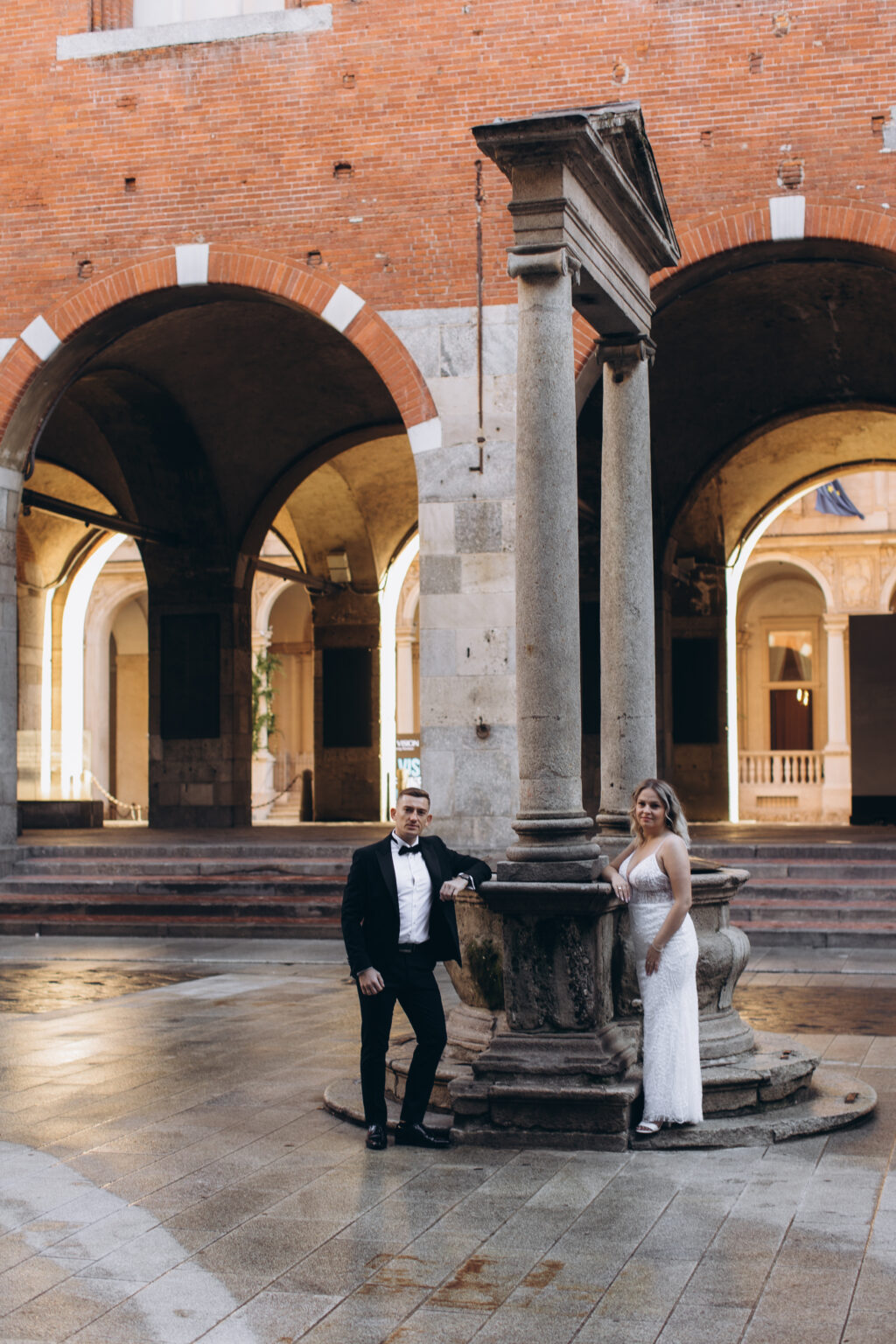 The couple lean on either side of an old stone well beneath brick arches in a historic courtyard in central Milan.