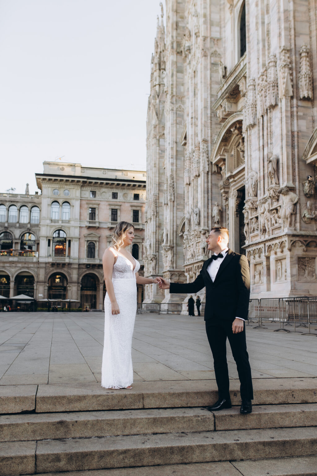He reaches for her hand across the low steps on Piazza del Duomo, the cathedral and the arcaded buildings of the square behind them.