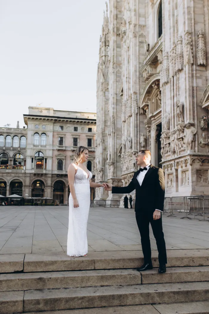 He reaches for her hand across the low steps on Piazza del Duomo, the cathedral and the arcaded buildings of the square behind them.
