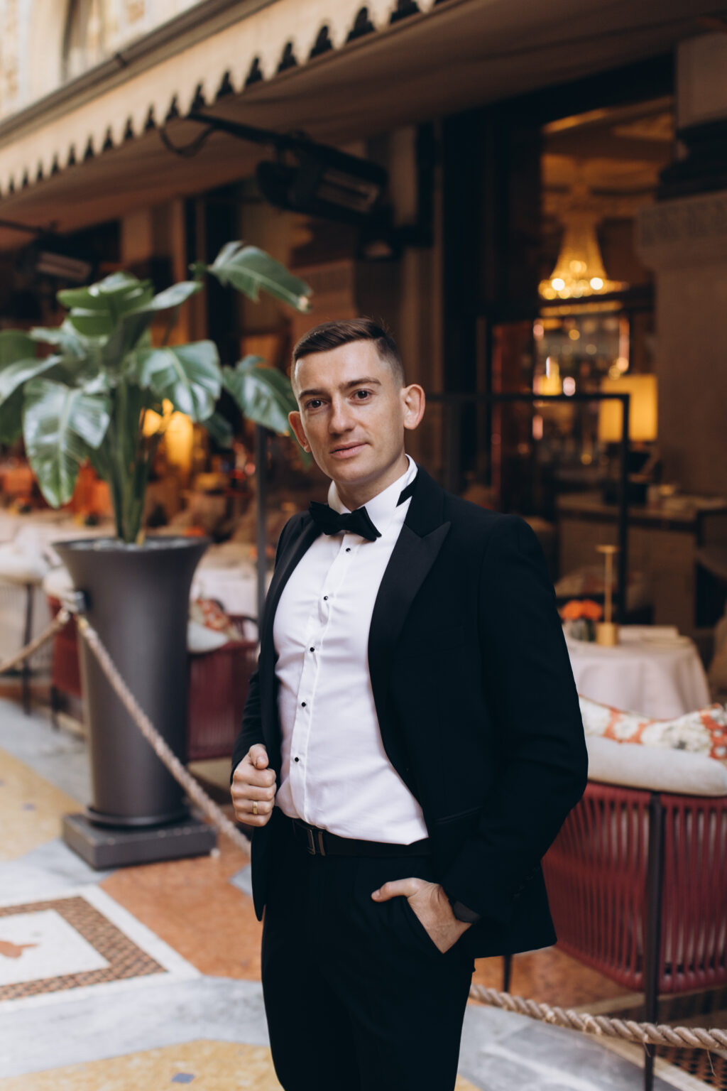 He stands in a black tuxedo before a café in the Galleria Vittorio Emanuele II, warm lights and a leafy plant behind him.