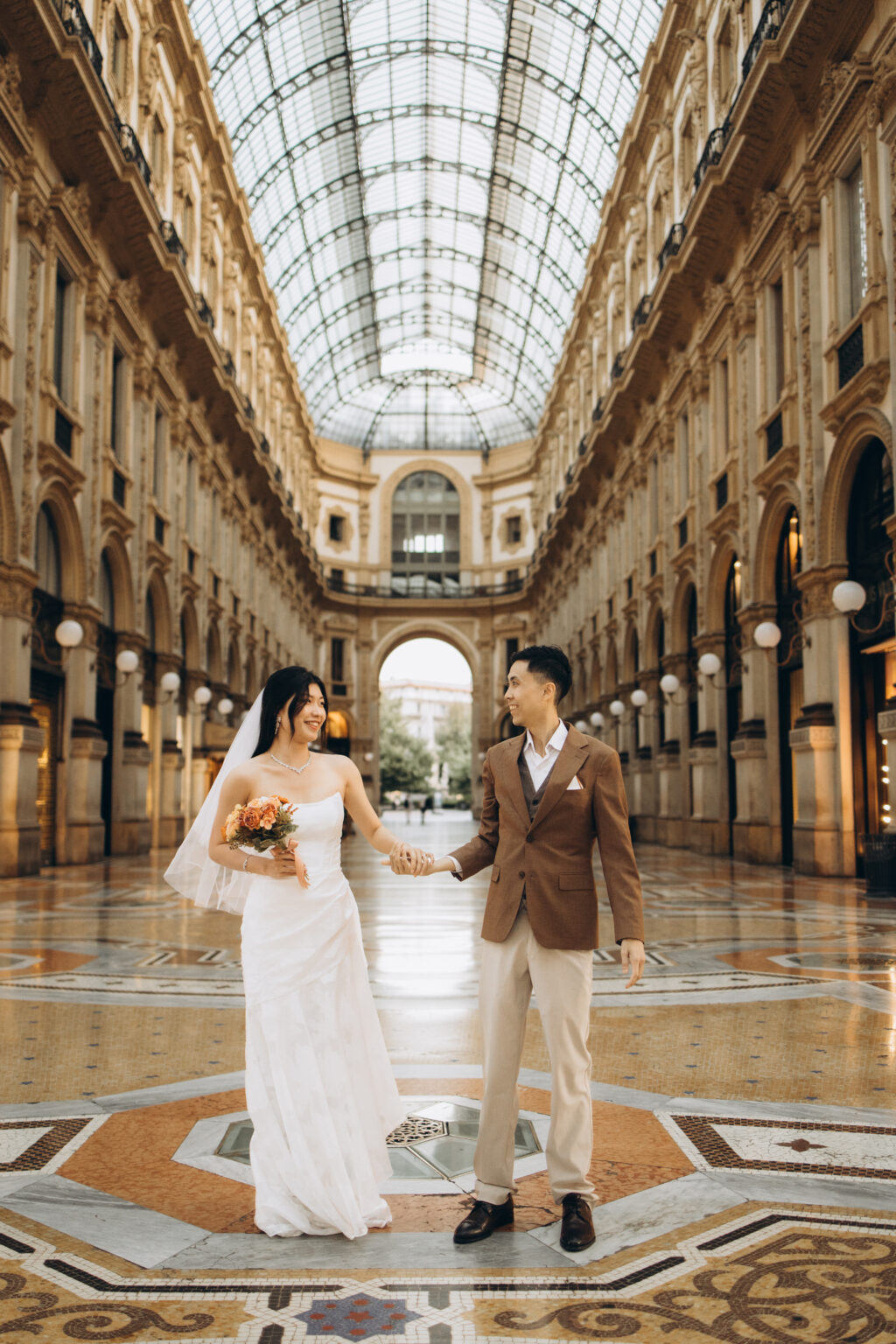 Bride-to-be in a white gown and veil holding hands with her groom-to-be at the center of the Galleria Vittorio Emanuele II in Milan