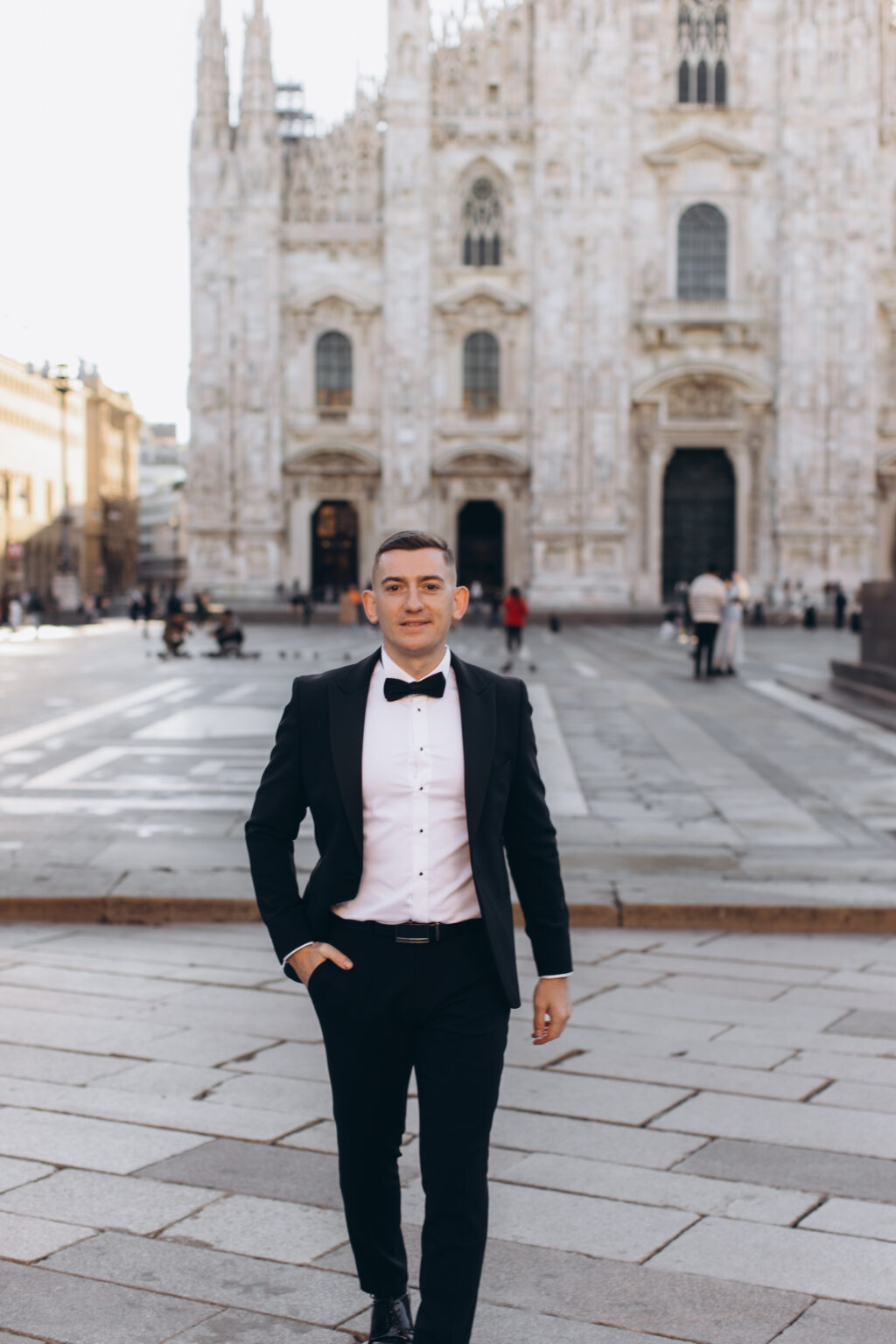 He walks toward the camera in a black tuxedo and bow tie on Piazza del Duomo, the marble facade of the Milan Cathedral behind him.