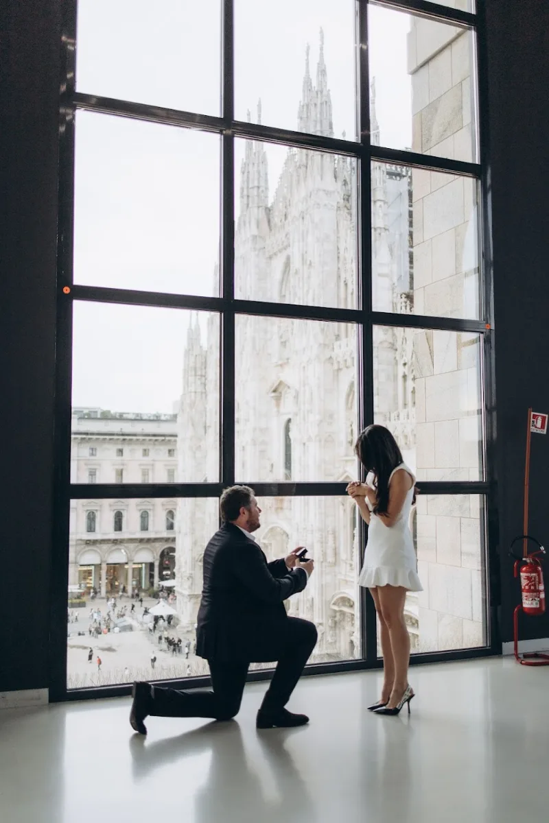 He kneels at a tall window above Piazza del Duomo as she turns, Milan