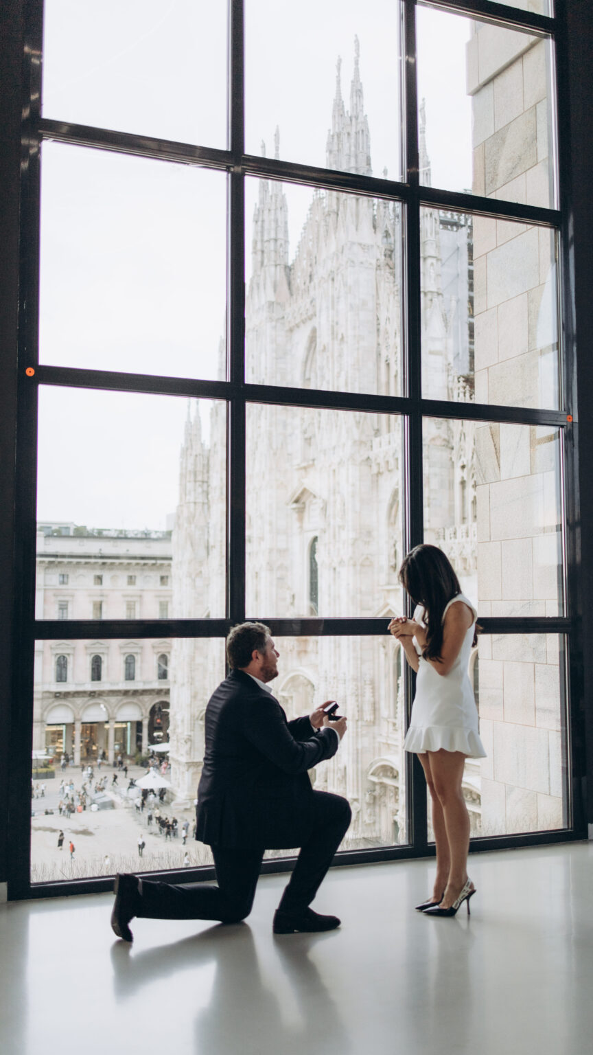 Man kneeling with an open ring box to propose beside the tall Museo del Novecento window that frames the Duomo di Milano in Milan