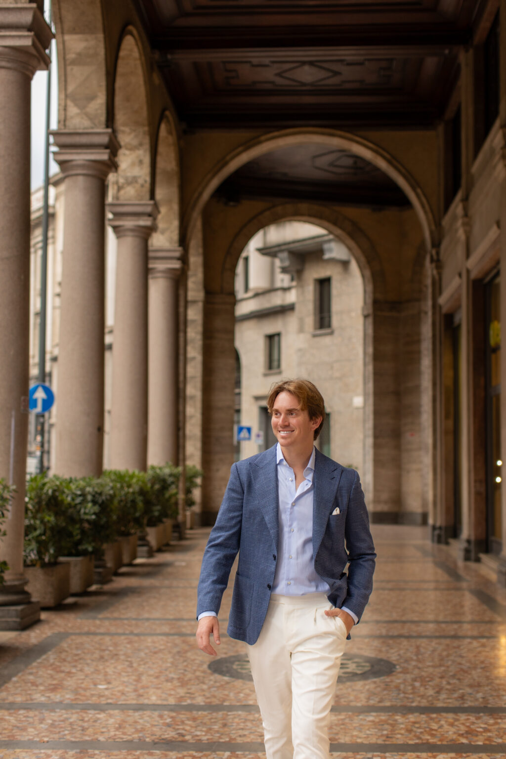 A man in a blue jacket walking toward the camera under the columned porticoes of a street in central Milan.