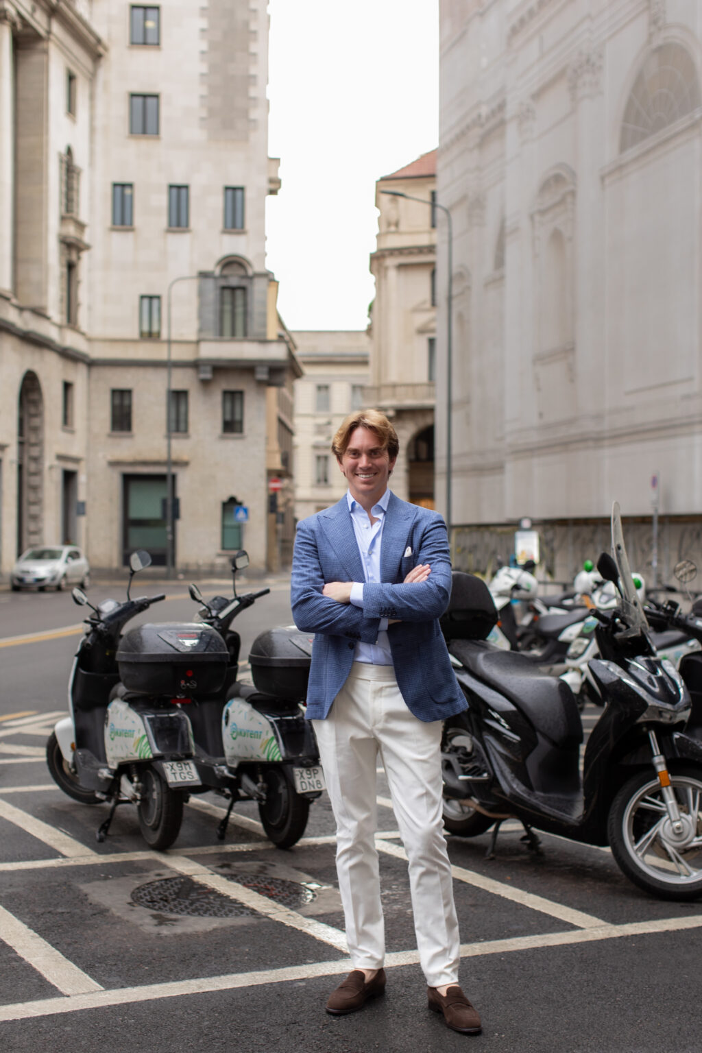 A man in a blue blazer standing with arms crossed on a Milan street beside parked scooters and mopeds.
