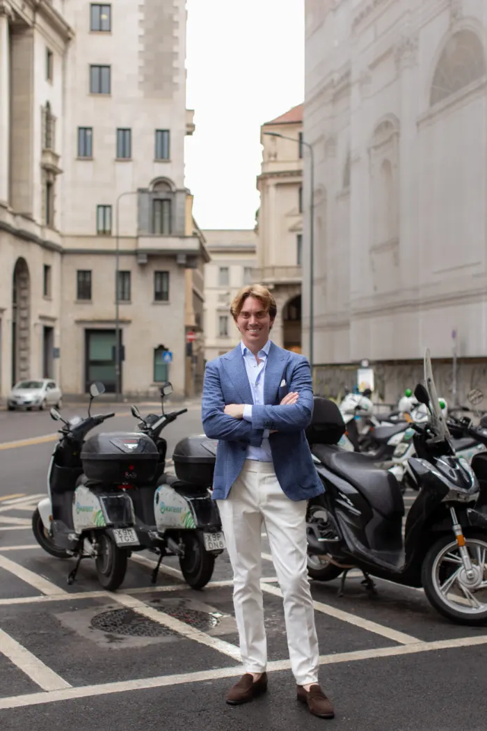 A man in a blue blazer standing with arms crossed on a Milan street beside parked scooters and mopeds.
