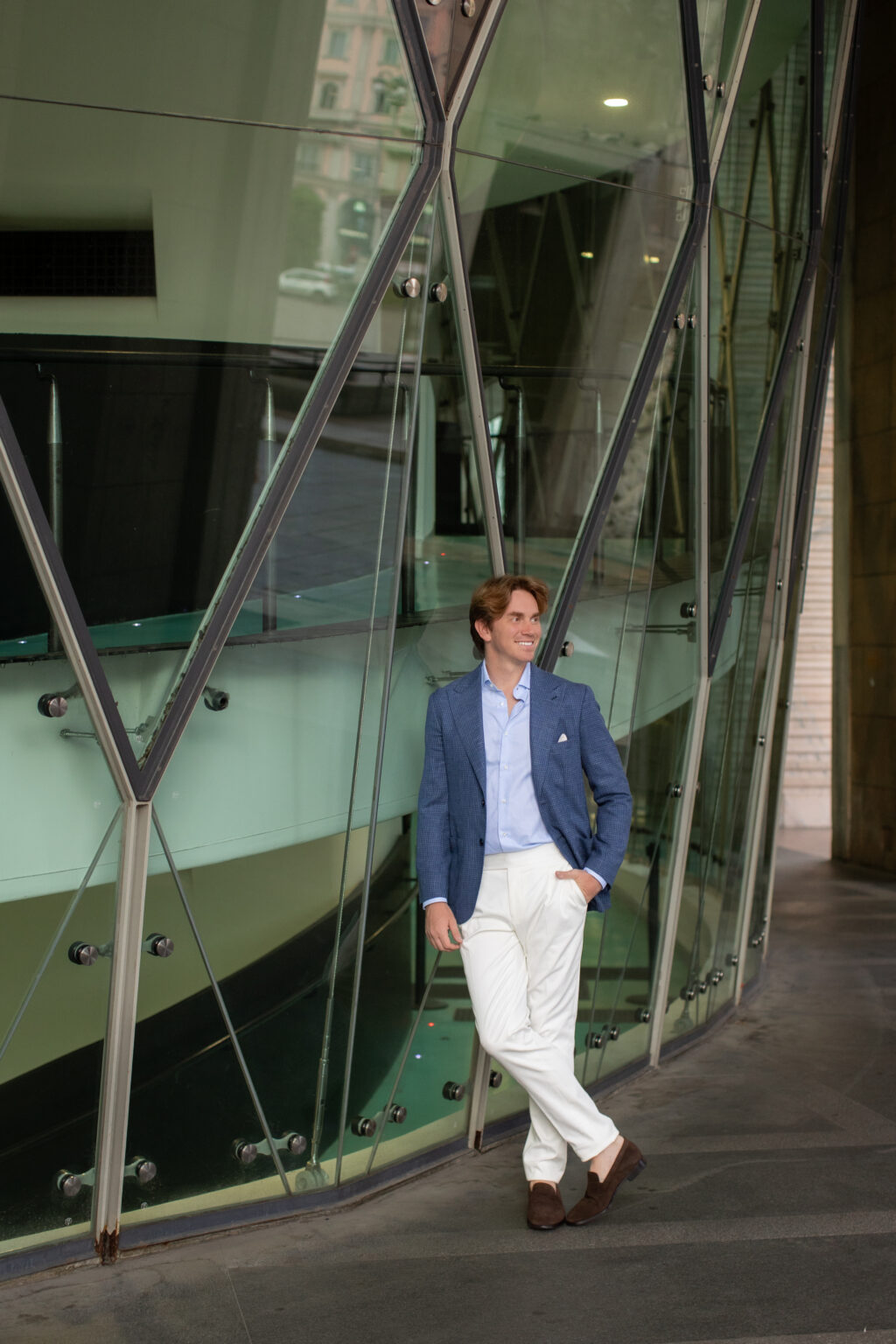 A full-length photo of a man in a blue blazer leaning against a curved modern glass building in Milan.