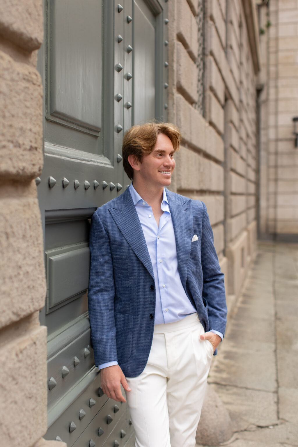 A man in a blue jacket leaning against a grey studded door along a quiet cobbled street in central Milan.